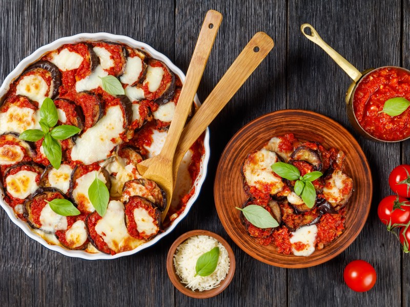 Parmigiana in einer Auflaufform, überbacken mit Mozzarella und Parmesan. Daneben eine Portion in einem Teller.