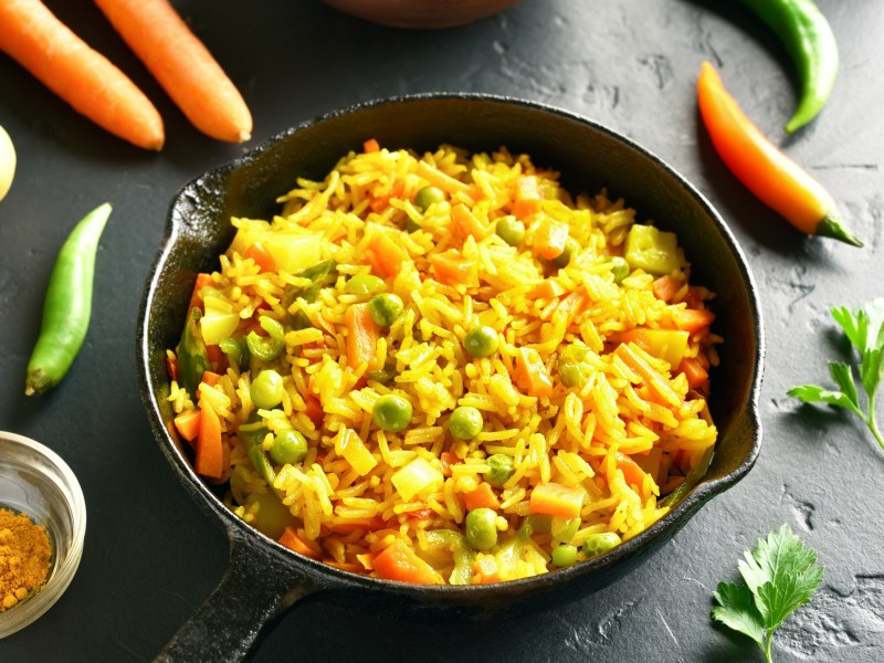 Reispfanne mit Gemüse wie Erbsen und Karotten, drumherum frische Zutaten.