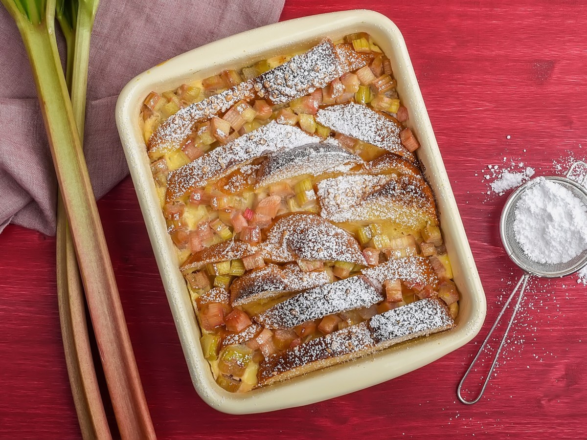 Auflaufform mit Brioche-Auflauf mit Rhabarber auf rotem Untergrund. Puderzucker und Rhabarber daneben.