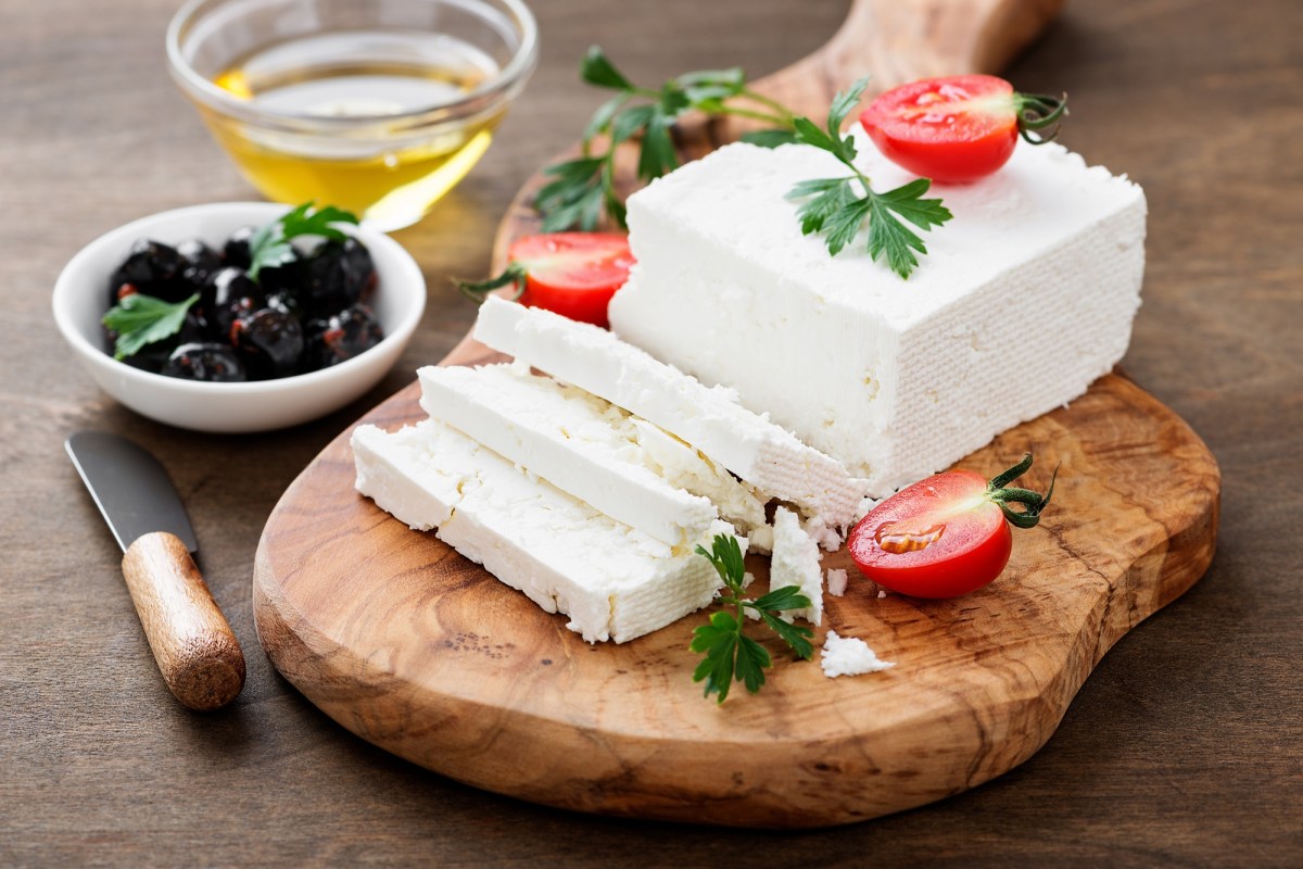 Unterschied Feta, Hirtenkäse und Schafskäse: Ein Stück Käse liegt auf einem Holzbrett. Garniert ist es mit TOmaten und Oliven.