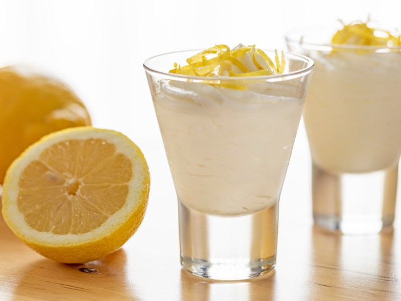 2 Gläser mit Zitronenmousse. Daneben liegt eine halbierte, frische Zitrone.