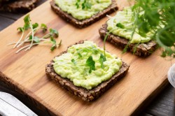 3 Scheiben Schwarzbrot mit Avocado-Dip mit Schmand bestrichen, darauf Kresse, die Brote liegen auf einem Brett.