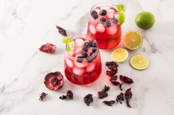 Zwei Gläser Blaubeer-Eistee, garniert mit frischen Blaubeeren. Drum herum liegen Hibiskusblüten und Limettenstücke.