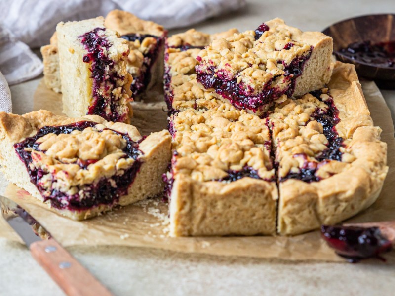 mehrere Stücke Streuselkuchen mit Marmelade auf einem Brett, daneben ein Holzlöffel dunkle Marmelade.