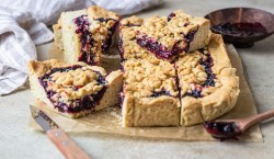 mehrere Stücke Streuselkuchen mit Marmelade auf einem Brett, daneben ein Holzlöffel dunkle Marmelade.