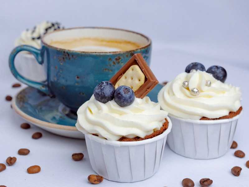 2 Cappuccino-Cupcakes dekoriert mit Schokolade, Heidelbeeren und Zuckerperlen, dahinter eine blaue Tassee Cappuccino.