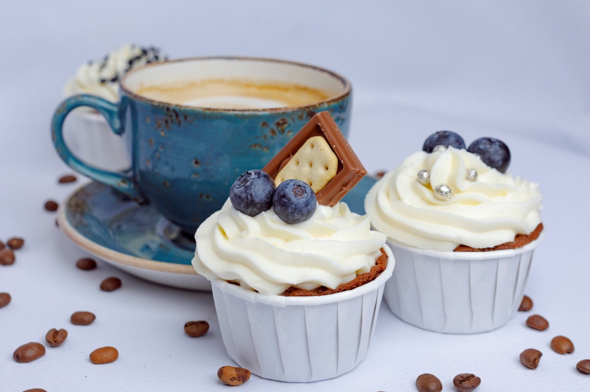 2 Cappuccino-Cupcakes dekoriert mit Schokolade, Heidelbeeren und Zuckerperlen, dahinter eine blaue Tassee Cappuccino.