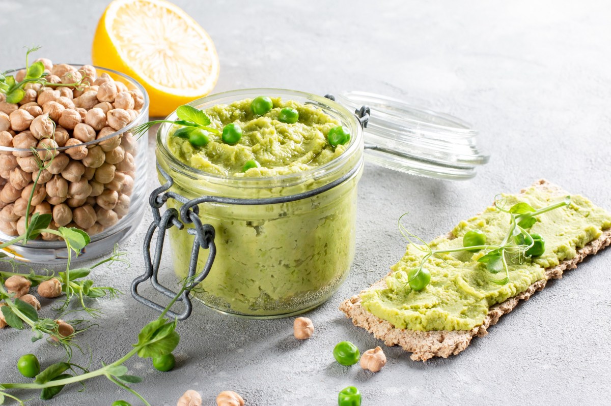 Erbsen-Hummus im Glas garniert mit Kresse, daneben Kichererbsen im Glas, eine halbe Zitrone und ein Knäckebrot mit Hummus bestrichen.