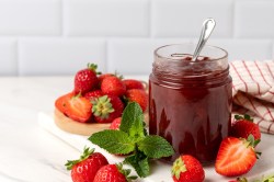 Ein Glas Erdbeer-Kokos-Marmelade mit einem Löffel darin, drumherum frische Erdbeeren.