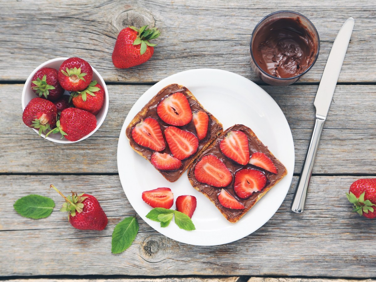 Erdbeer-Nutella-Toasts auf einem weoßen Teller. Daneben stehen ein Glas Nutella und es liegen einige Erdbeeren bereit.