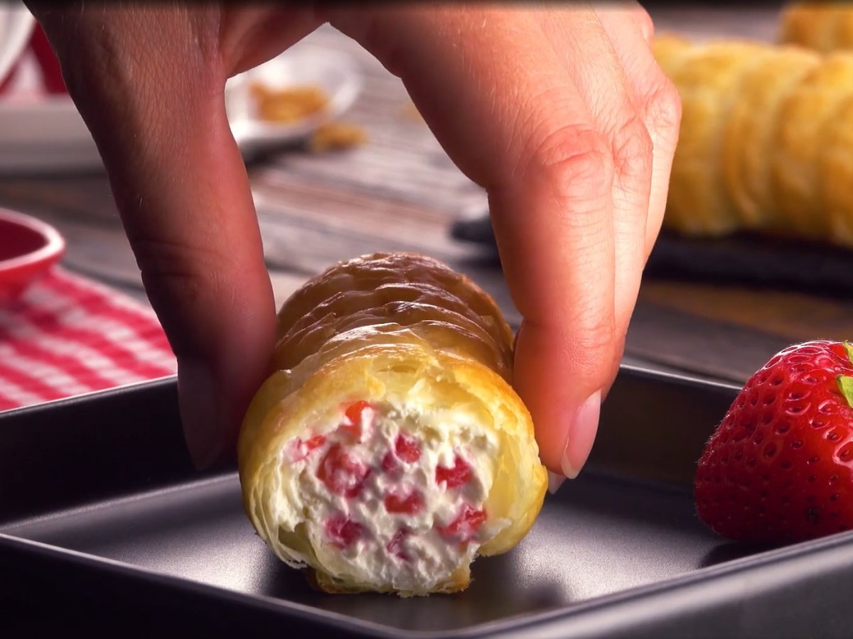 Ein Erdbeer-Röllchen wird auf einen dunklen Dessertteller gelegt, daneben eine Erdbeere.