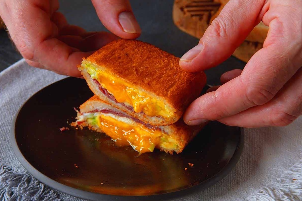 Ein gebackenes Sandwich auf einem Teller, das halbiert wurde und von zwei Händen gehalten wird.