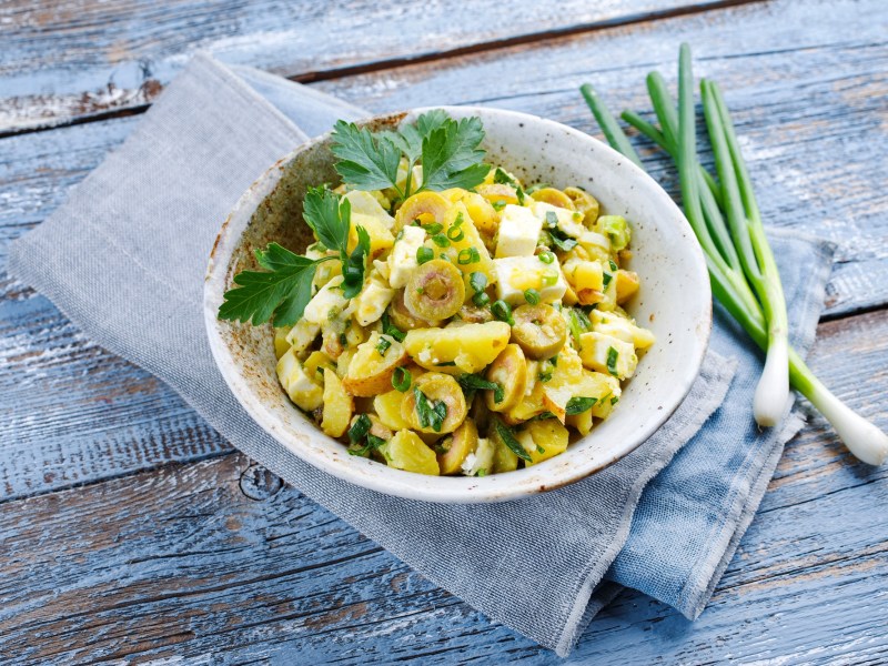 Griechischer Kartoffelsalat mit Oliven und Feta sowie Dill und Frühlingszwiebeln in einer Schüssel, die auf einer Stoffserviette liegt, daneben frische Frühlingszwiebeln.