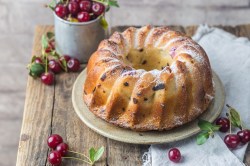 Ein Gugelhupf mit Kirschen auf einem Holztisch mit frischen Kirschen.