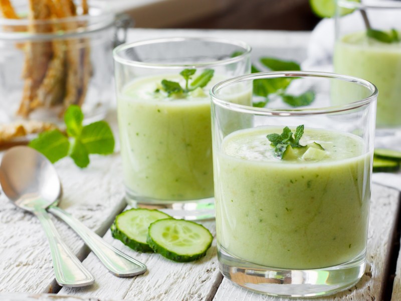 2 Gläser Gurken-Gazpacho garniert mit Minze, daneben zwei Löfel und Gurkenscheiben.