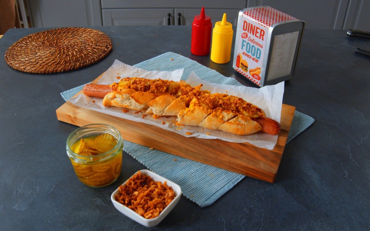 Ein Hotdog-Baguette auf einem Brett. Daneben stehen Ketchup, Senf, weitere Zutaten und ein Serviettenhalter.