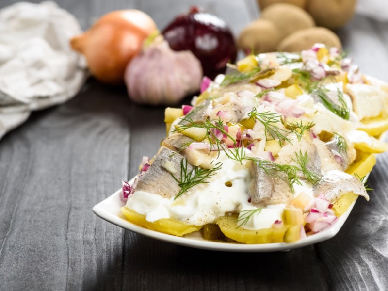 Ein Teller mit Kartoffelsalat mit Hering. Im Hintergrund liegen frische Zutaten.