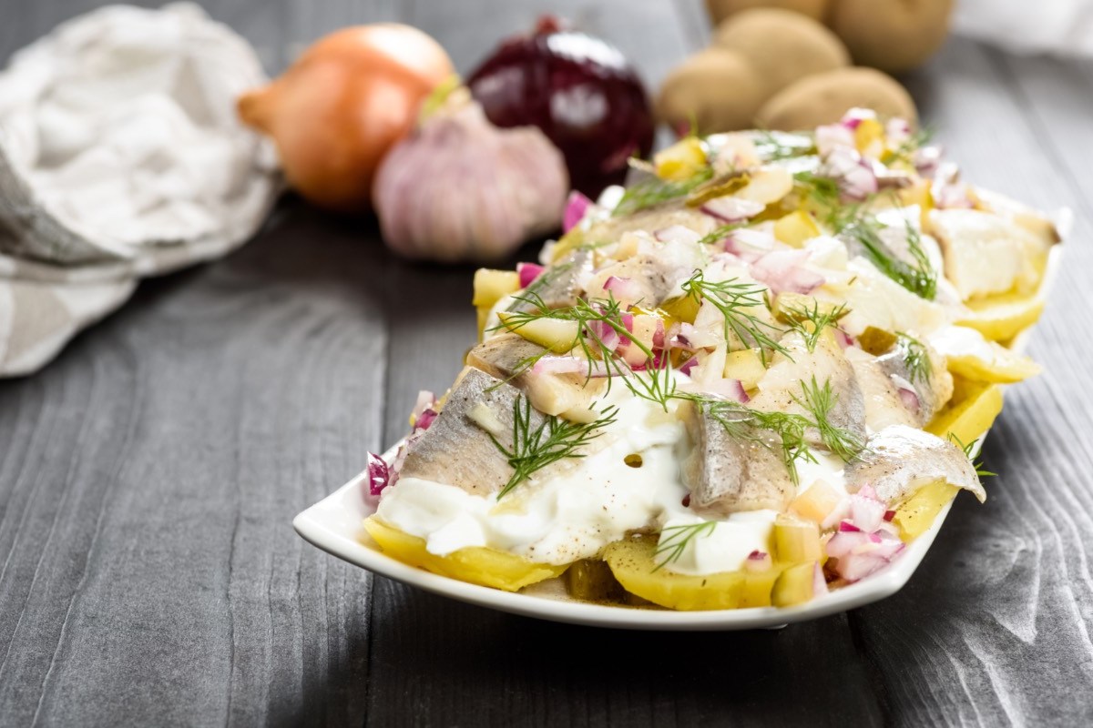 Ein Teller mit Kartoffelsalat mit Hering. Im Hintergrund liegen frische Zutaten.