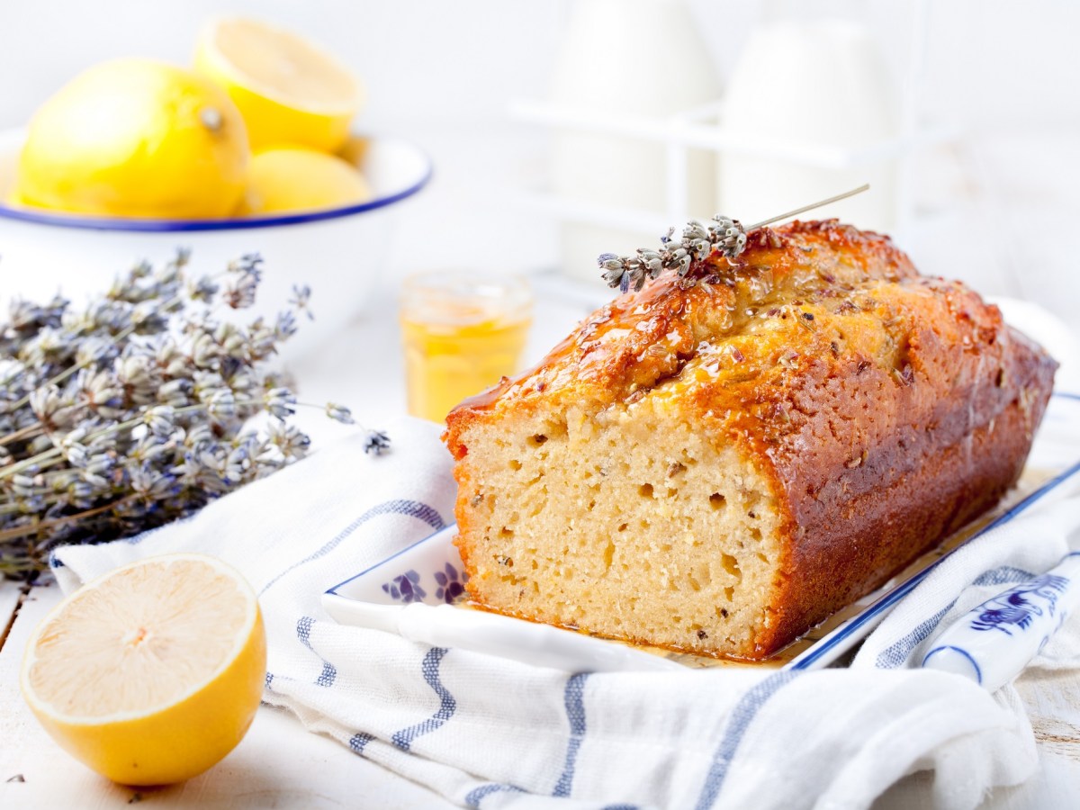 ein angeschnittener franzÃ¶sischer Lavendelkuchen auf einem karierten Geschirrtuch, daneben Lavendel und Zitronen.