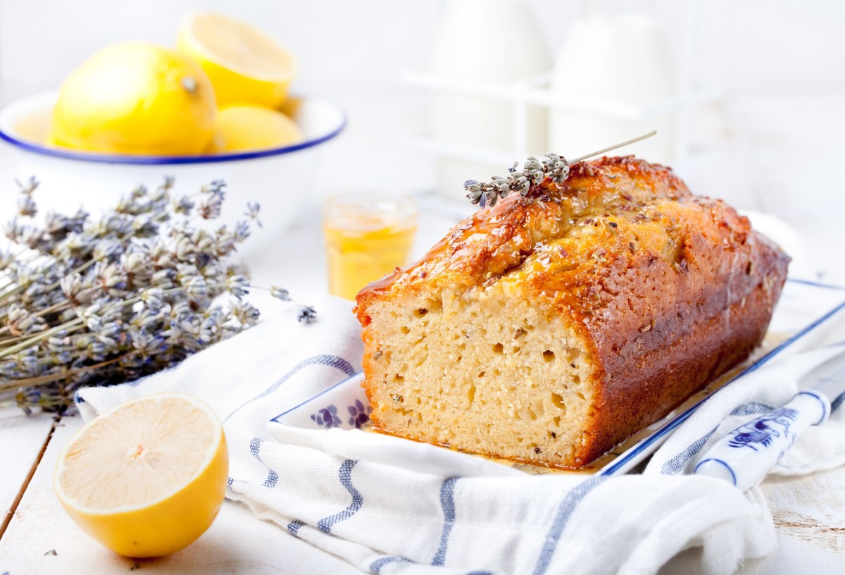 ein angeschnittener französischer Lavendelkuchen auf einem karierten Geschirrtuch, daneben Lavendel und Zitronen.