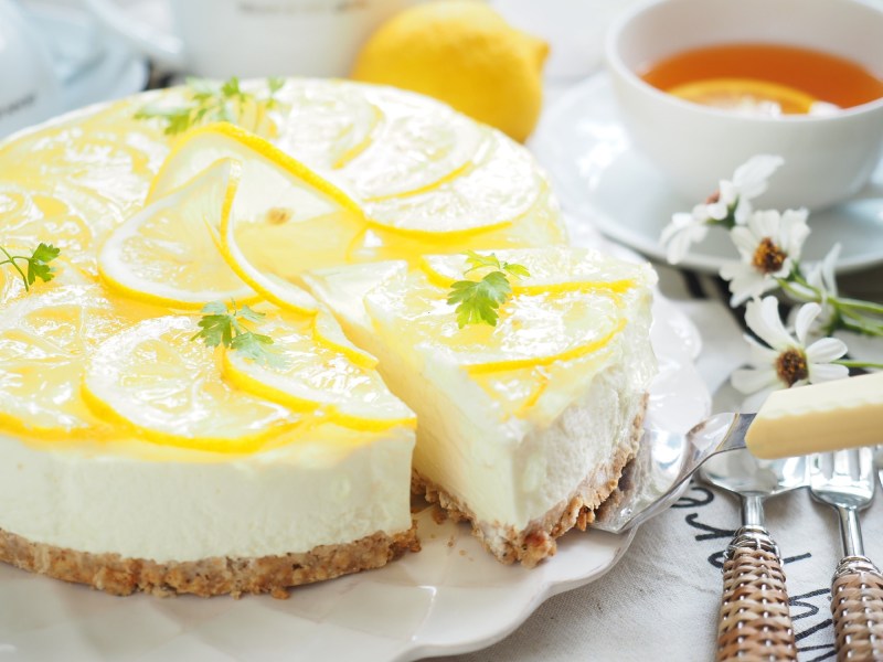 Ein Limoncello-Cheesecake, aus dem ein Stück mit einem Tortenheber entnommen wird. Der Kuchen ist garniert mit Zitronenscheiben, daneben steht eine Teetasse. Nahaufnahme.