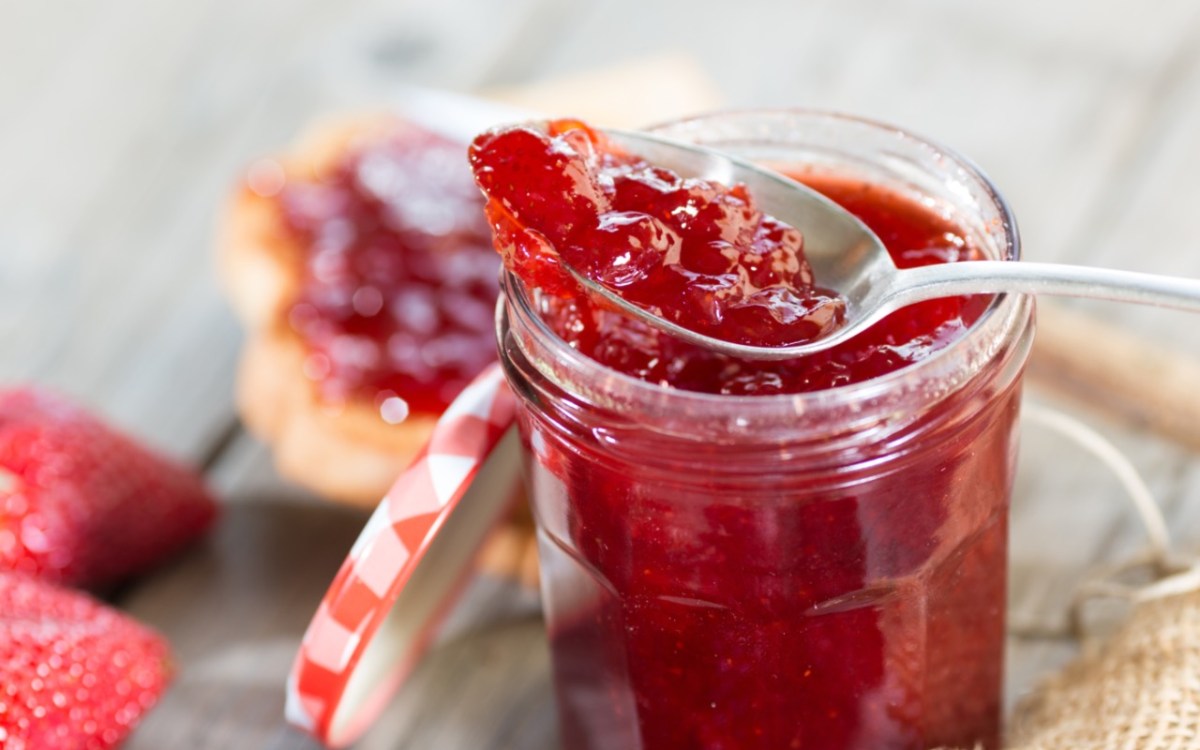 Marmelade einfrieren: ein Glas Erdbeermarmelade mit einem Löffel darin. Im Hintergrund Brötchen mit Marmelade darauf.
