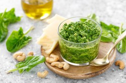 Ein Glas Spinatpesto auf einem runden Holzteller, drumherum frischer Spinat, Cashews und außerdem ein Glas Olivenöl.