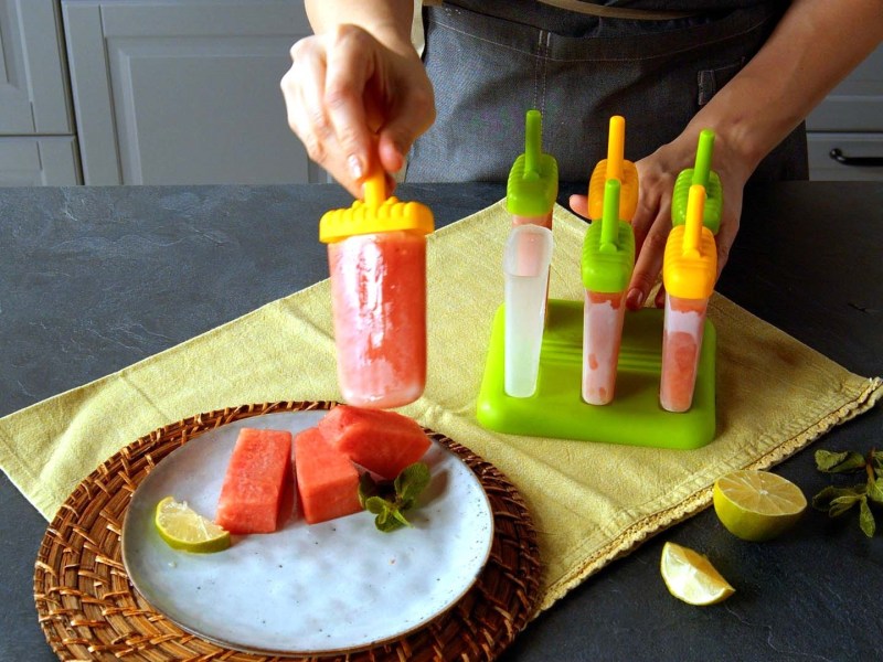 ein selbst gemachtes Wassermelonen-Eis wird von einer Frauenhand gehalten, daneben eine Schüssel mit Wassermelonenstückchen und mehr Eis.