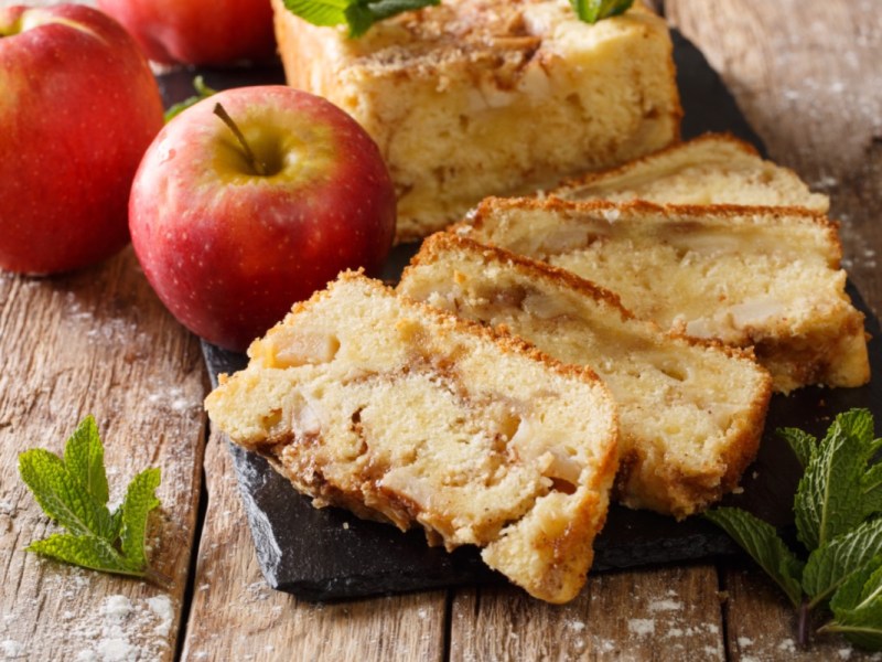 Mehrere Stücke Apfelbrot, die gefächert vor dem restlichen Kuchen liegen. Daneben zwei Äpfel.