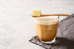 Bulletproof Coffee in einem Glas, darüber ein Löffel mit einem Stück Butter.