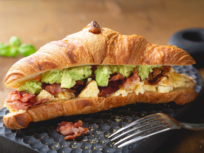 Ein Croissant mit Rührei Avocado und Speck auf einer Schieferplatte, davor eine Gabel.