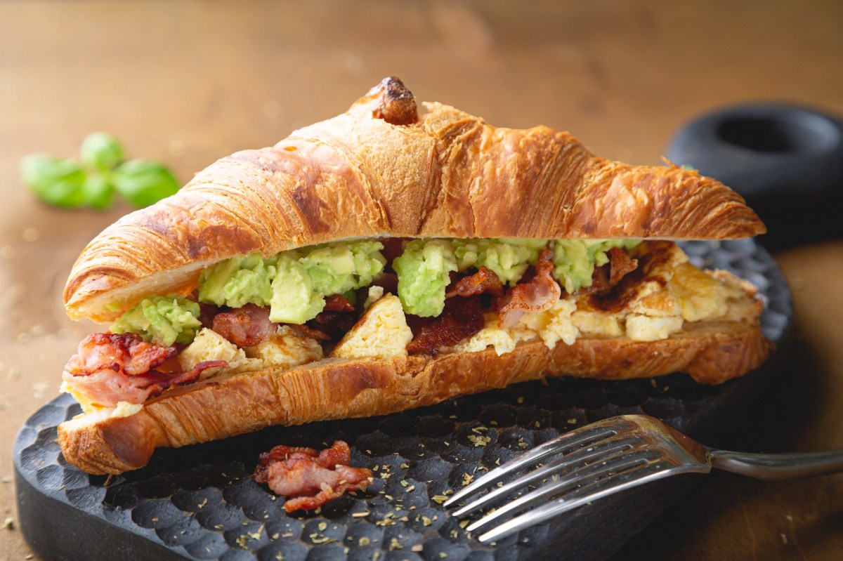 Ein Croissant mit Rührei Avocado und Speck auf einer Schieferplatte, davor eine Gabel.