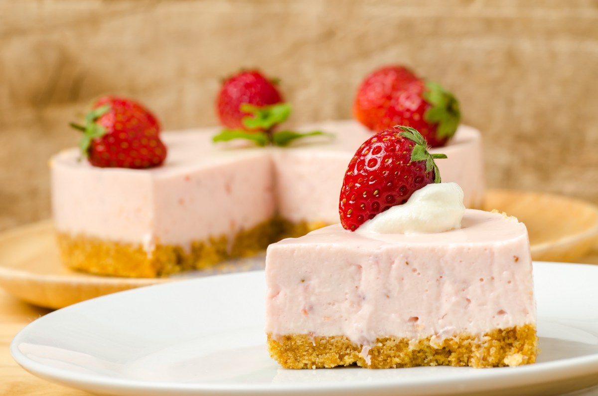 Ein StÃ¼ck Erdbeermousse-Kuchen, garniert mit einer Erdbeere, auf einem Teller, im Hintergrund der angeschnittene Kuchen.