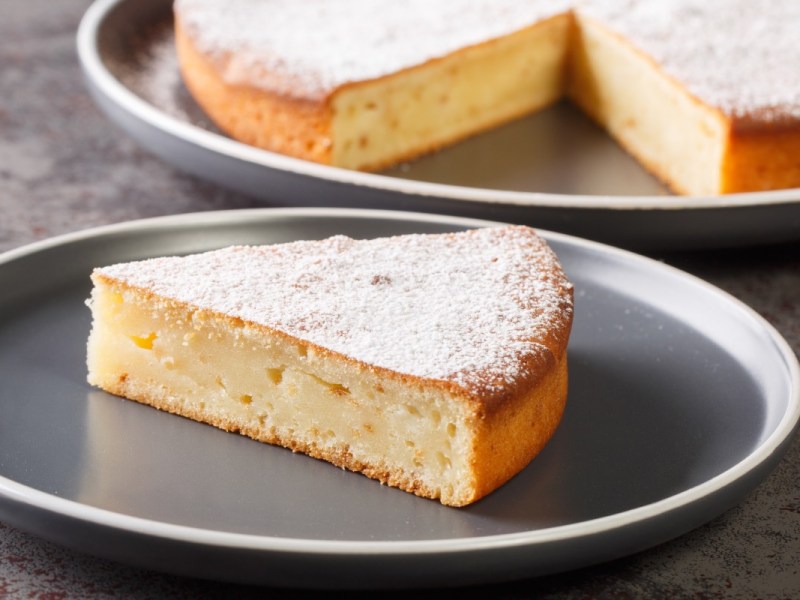 Ein Stück Joghurtkuchen mit 3 Zutaten auf einem Teller. Im Hintergrund der restliche Kuchen.