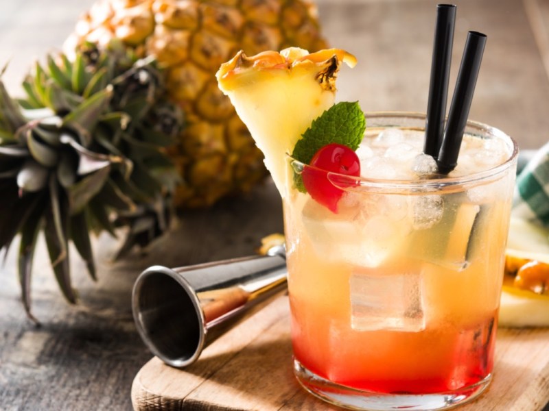 Ein Glas Mai Tai mit zwei Strohhalmen. Dahinter liegen Cocktailzubehör und eine Ananas.