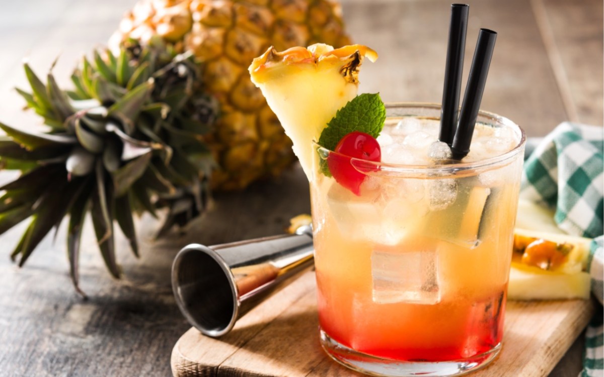 Ein Glas Mai Tai mit zwei Strohhalmen. Dahinter liegen Cocktailzubehör und eine Ananas.