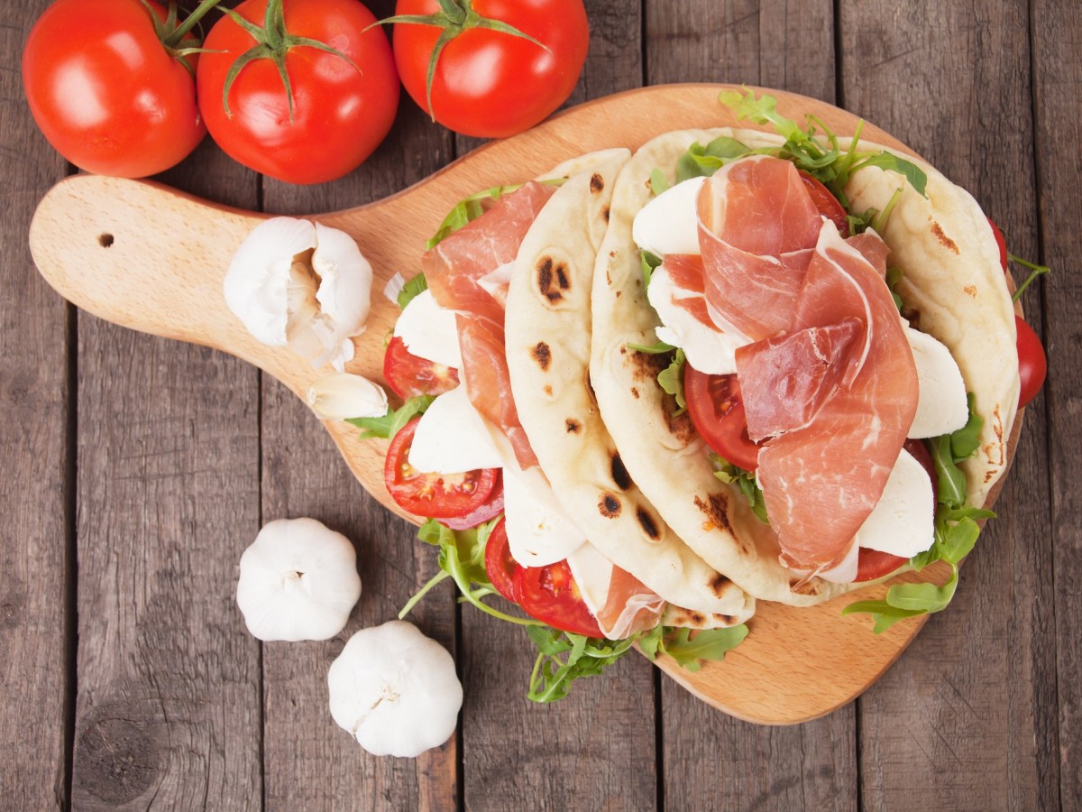 Piadine auf einem Holzbrett. Daneben liegen TOmaten und Knoblauchknollen.