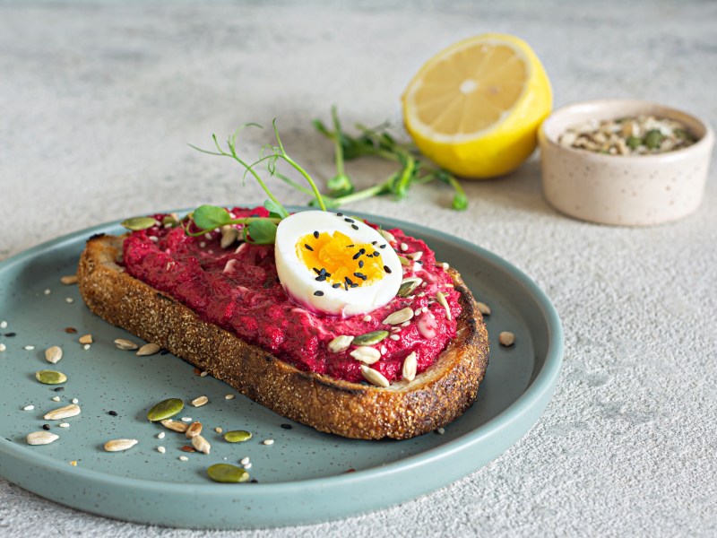 Eine Scheibe Brot mit Rote-Bete-Aufstrich mit Frischkäse und Ei auf einem Teller, garniert mit Kernen, im Hintergrund eine halbe Zitrone und eine Schale Kerne.