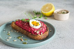 Eine Scheibe Brot mit Rote-Bete-Aufstrich mit Frischkäse und Ei auf einem Teller, garniert mit Kernen, im Hintergrund eine halbe Zitrone und eine Schale Kerne.