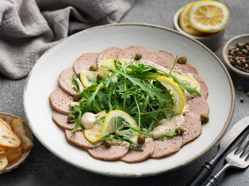 Vitello Tonnato auf einem weißen Teller, garniert mit Rucola und Zitrone.