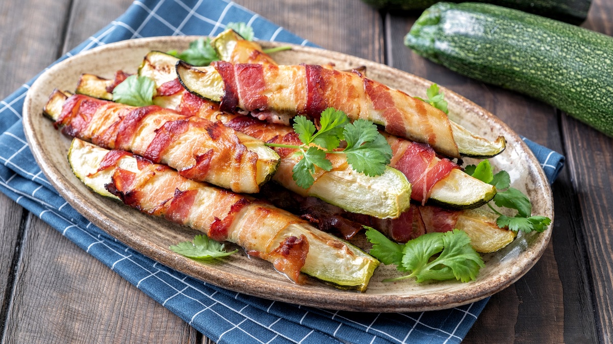 Zucchini im Speckmantel auf einem ovalen Teller, daneben eine Zucchini.