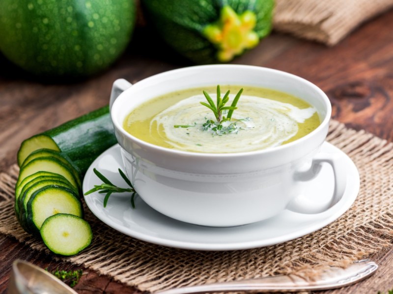 Eine Schüssel mit Zucchini-Suppe. Daneben liegt eine aufgeschnittene Zucchini.