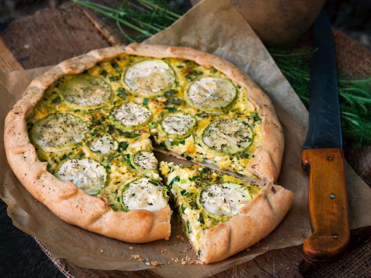 Zucchini-Tarte mit Ziegenkäse auf einem Backpapier, das auf einem Holzbrett liegt. Ein Stück wurde bereits abgeschnitten.