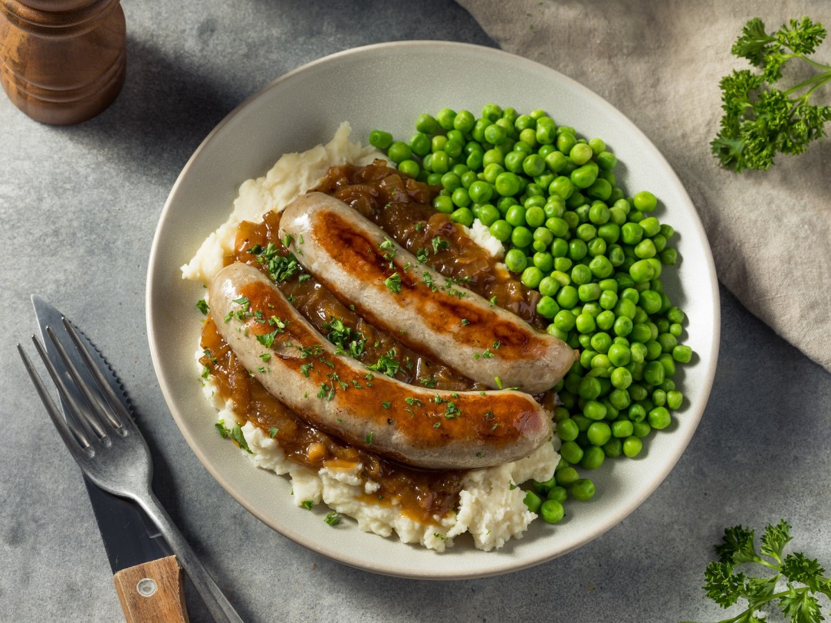 Bangers and Mash: ein Teller mit zwei Bratwürsten, Kartoffelbrei, Erbsen und brauner Soße, daneben Messer und Gabel, Draufsicht.