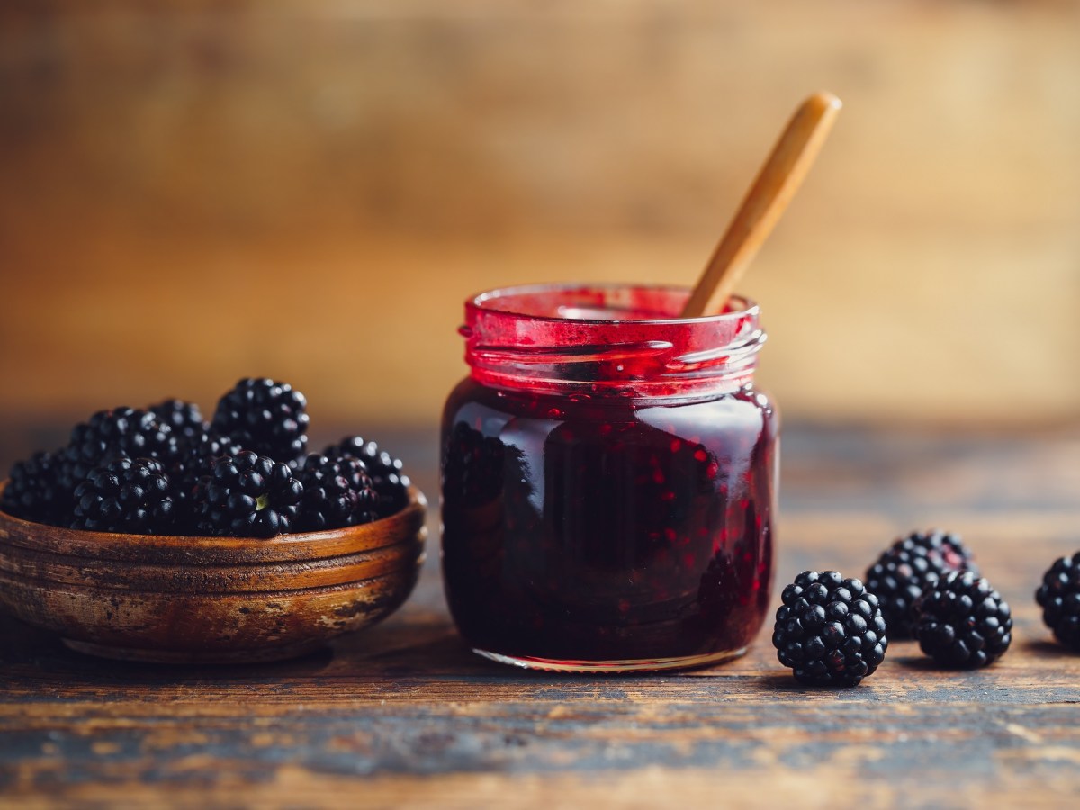 Brombeermarmelade in einem Glas, das auf einem Holztisch steht. Daneben liegen ganze Brombeeren. In der Marmelade steckt ein Holzlöffel.