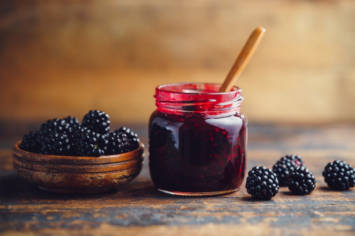 Brombeermarmelade in einem Glas, das auf einem Holztisch steht. Daneben liegen ganze Brombeeren. In der Marmelade steckt ein Holzlöffel.