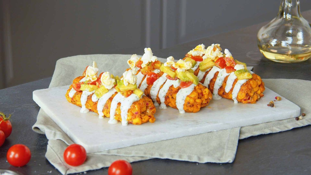 3 Käse-Mais-Kroketten auf einem Teller, garniert mit Sour Cream, Tomaten-Avocado-Salat und Popcorm.