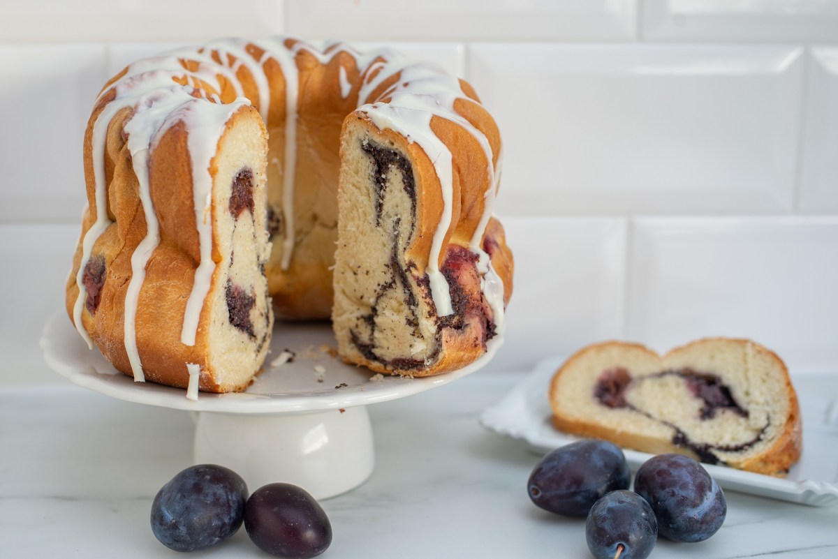 Ein angeschnittener Marmorkuchen mit Pflaumen in Gugelhupf-Form, daneben ein Teller mit einem Stück sowie frische Pflaumen.