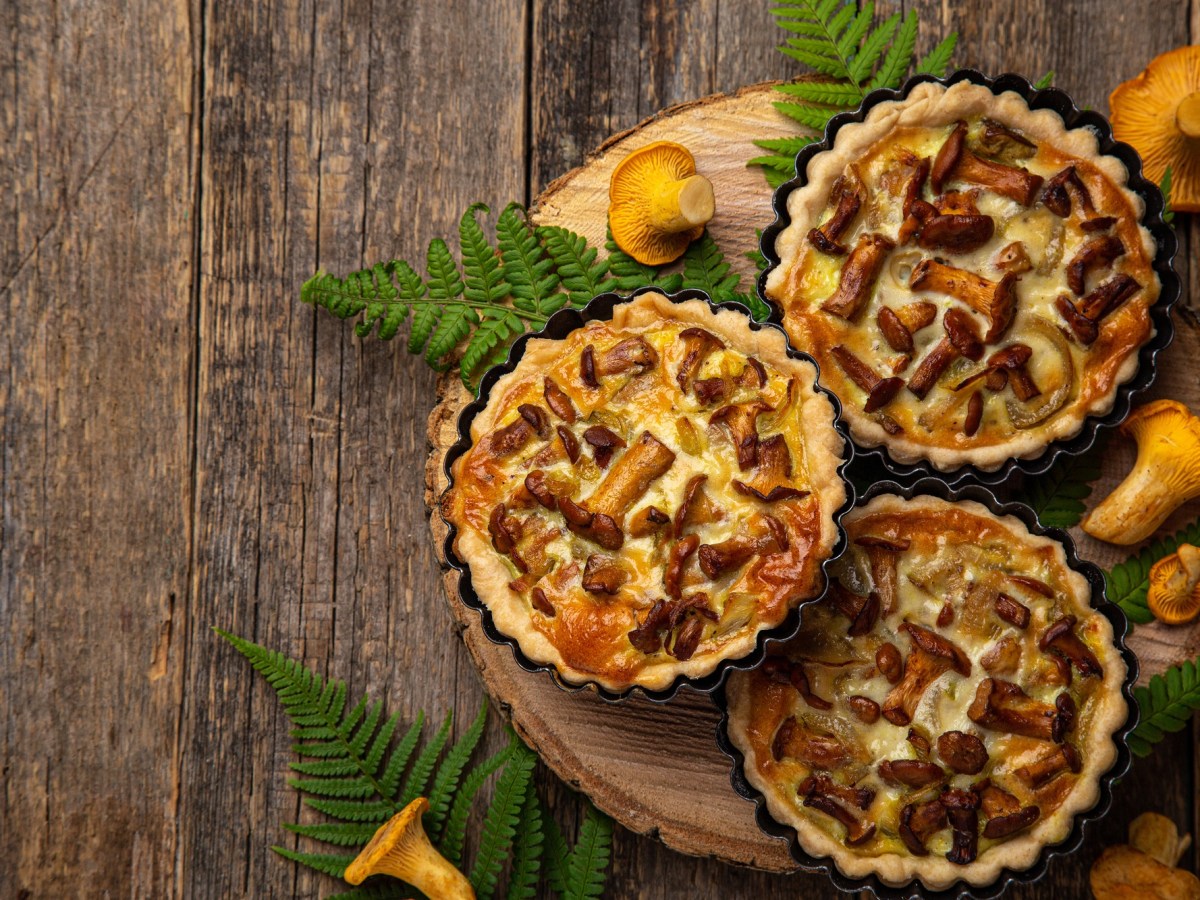 Drei Pfifferling-Tartelettes auf einem Holzbrett. Drum herum liegen Pfifferlinge und Farnblätter.