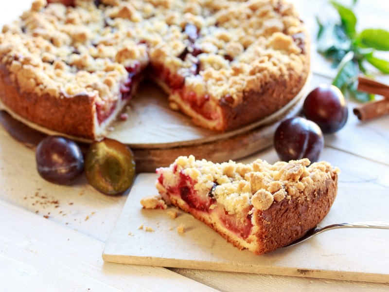 angeschnittener Pflaumenkuchen mit Zimtstreuseln, ein Kuchenstück liegt auf einem Brett davor.